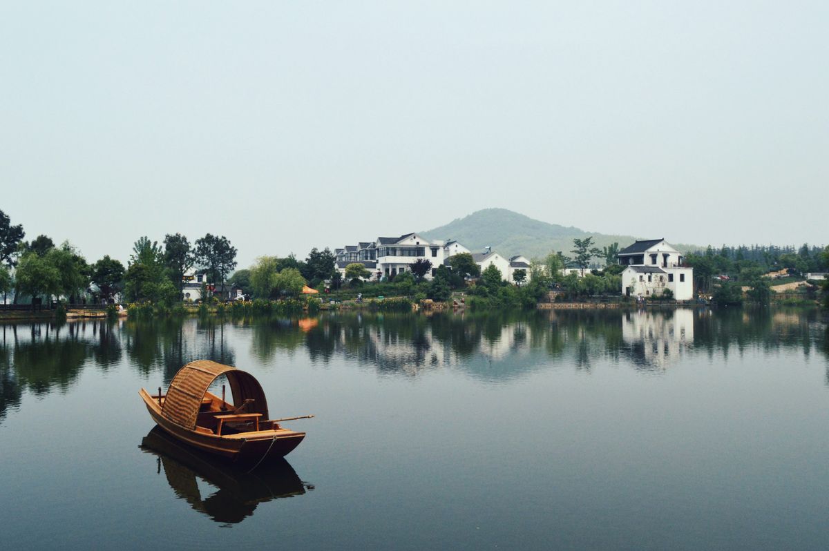 Brown wooden boat on body of water overlooking houses by the shore at daytime. Photo by Jennifer Chen.